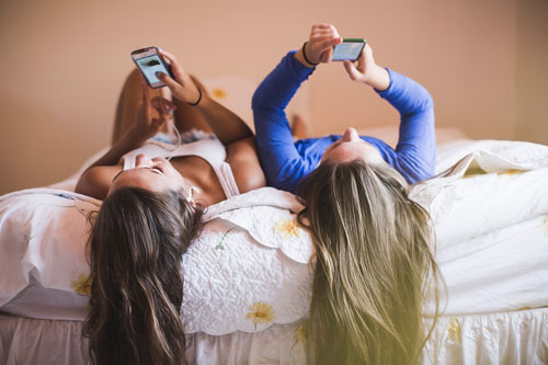 Två tjejer ligger och surfar på mobilen i en säng